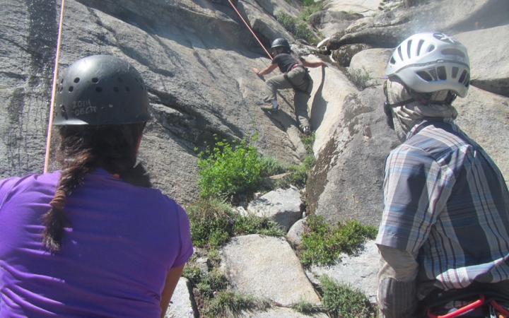two people wearing helmets look on as another person rock climbs on an outward bound course for bipoc